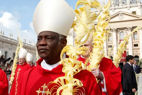 Reuters A mid-crop of Cardinal Peter Turkson looking solemnly ahead while wearing traditional religious robes along with others outside St. Peter's Basilica in April 2019.